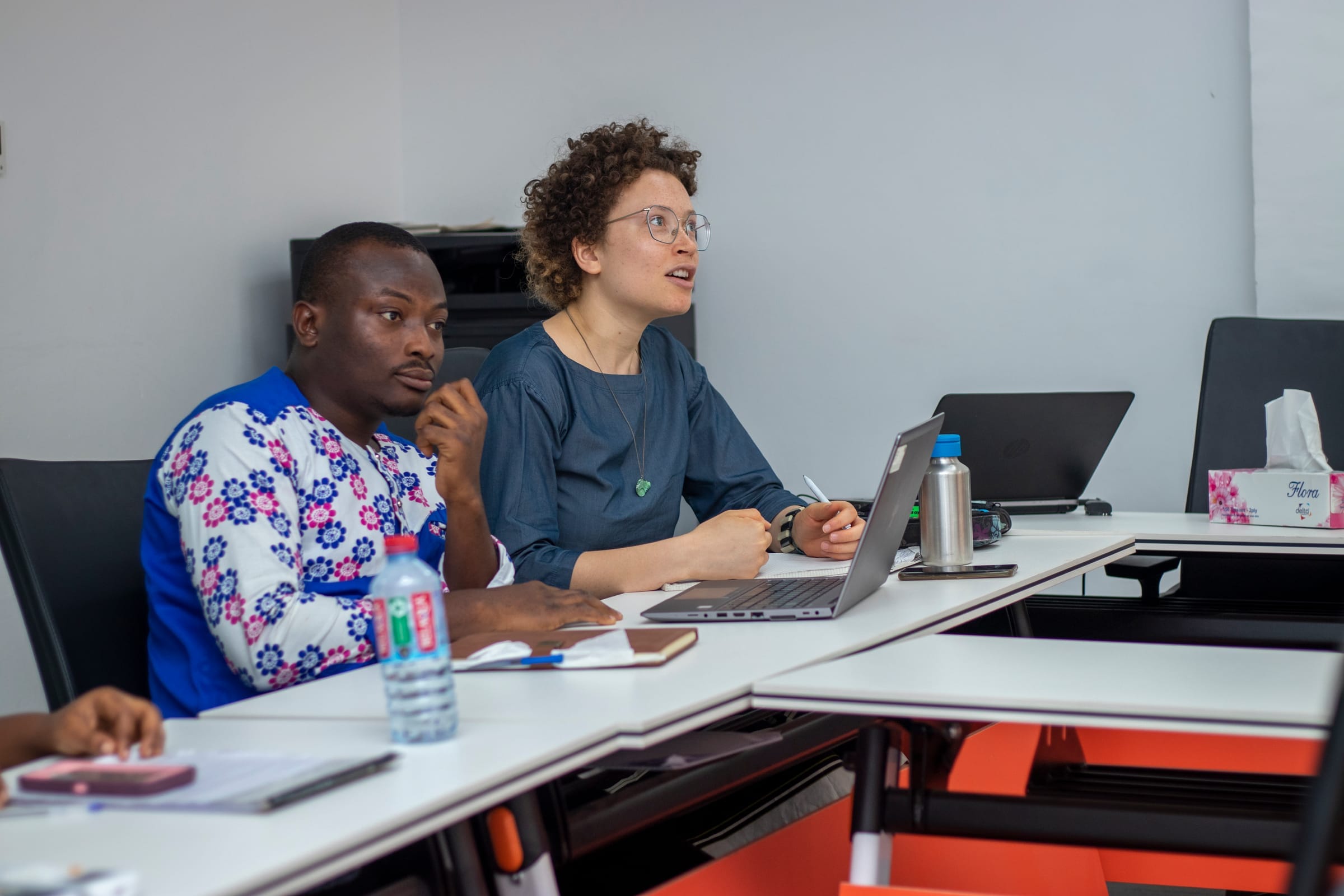 Two professionals at a desk in a bright room, focused on a speaker; laptop and notes suggest a workshop or seminar.