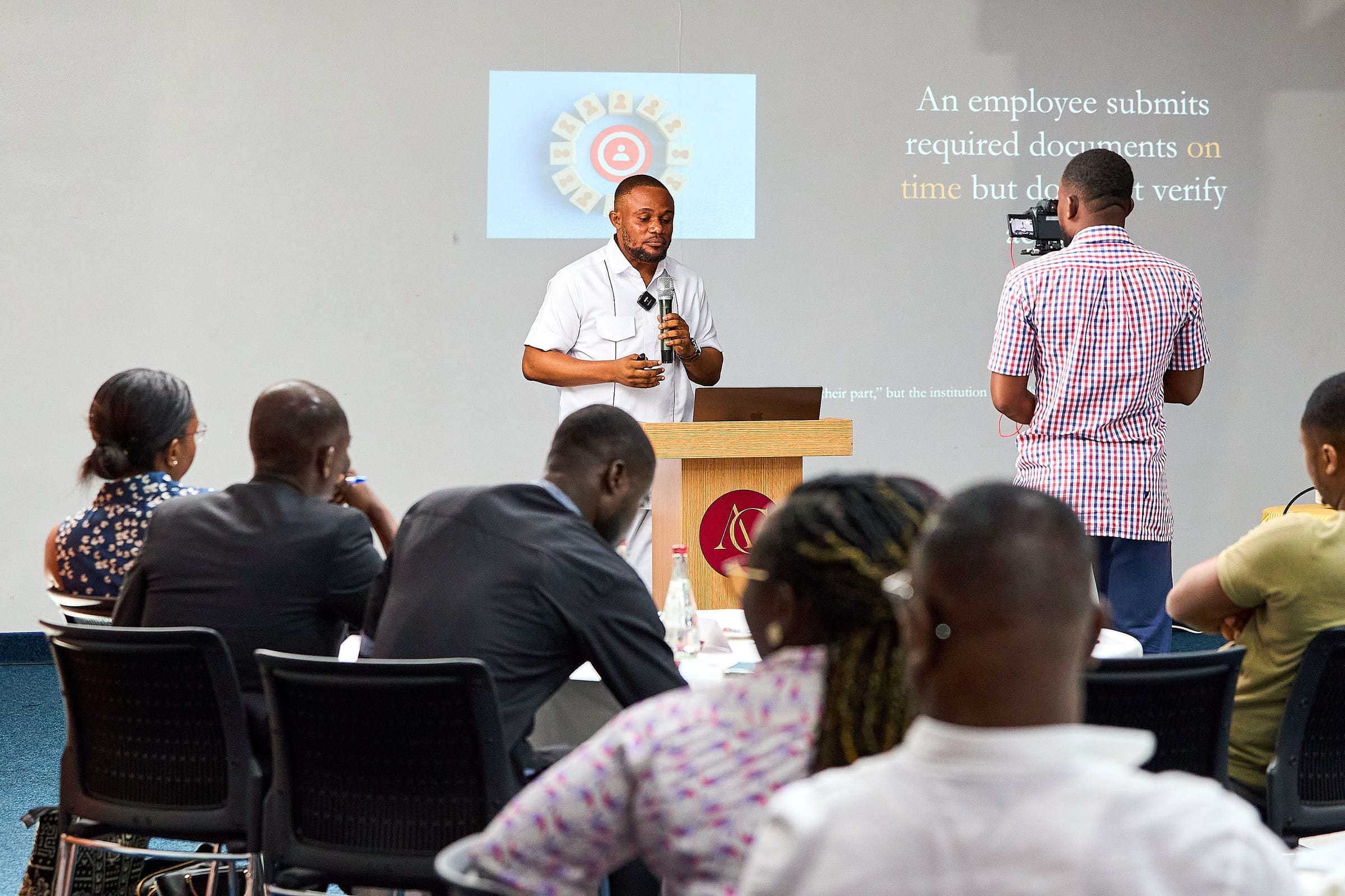 Presenter at a lectern speaking to an audience with a projected slide behind him; a videographer films the session.