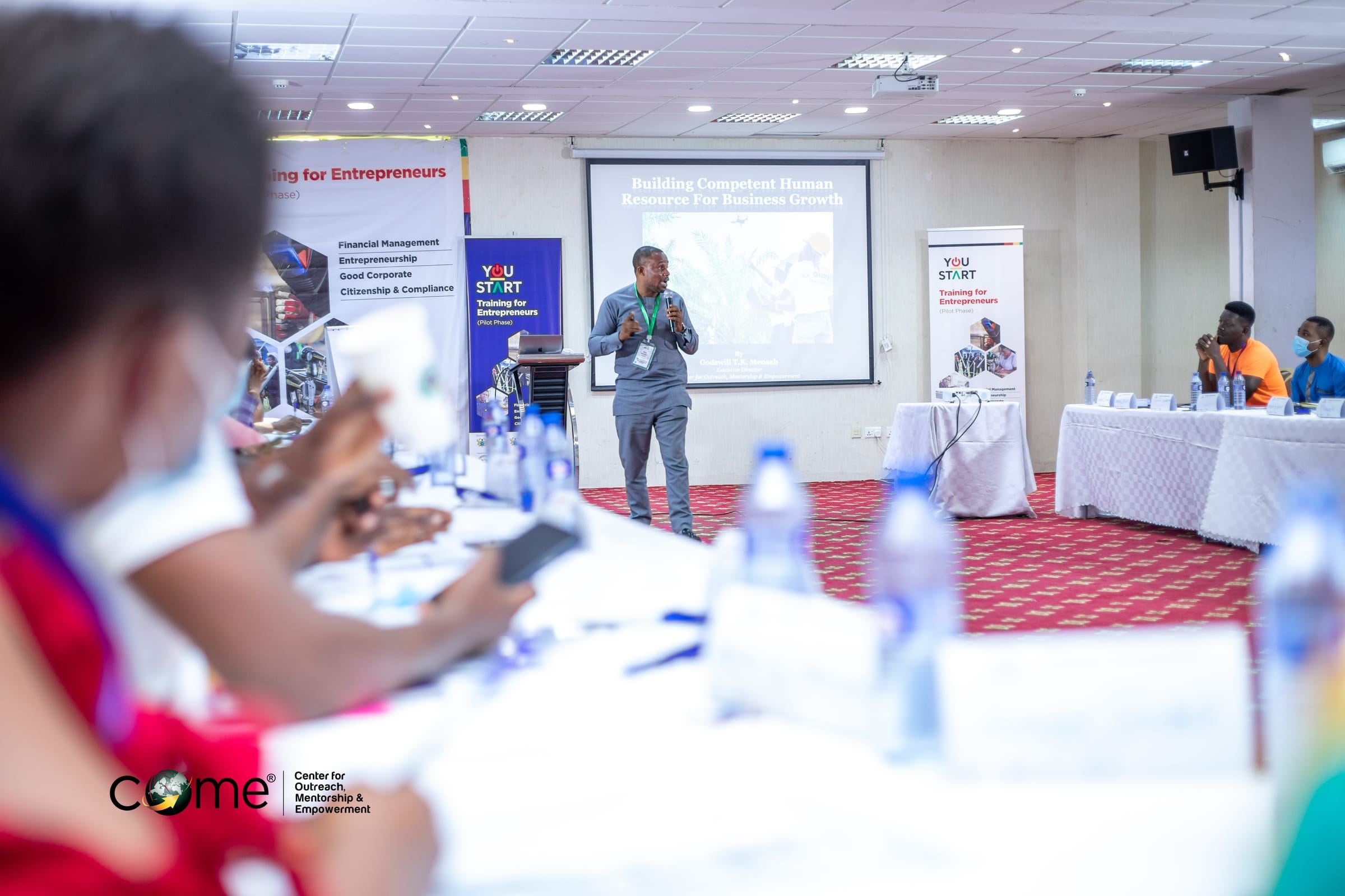 COME training session: facilitator speaking to participants in a workshop on building competent human resources and business growth, with COME branding visible.